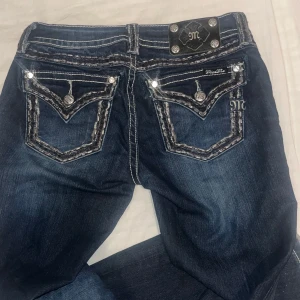 Mörkblå jeans från Miss Me - Snygga mörkblå jeans från Miss Me med dekorativa bakfickor och gnistrande detaljer. Jeansen har en låg midja och Bootcut passform, perfekt för en trendig look. Midjemått:37 Innerbenslängd: 84. Pris kan diskuteras vid snabb affär.