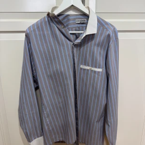 Randig skjorta från Melka Goldwin - Snygg randig skjorta från Melka Goldwin i blått och brun. Skjortan har en klassisk krage och en bröstficka med knappdetalj. Perfekt för en stilren look.