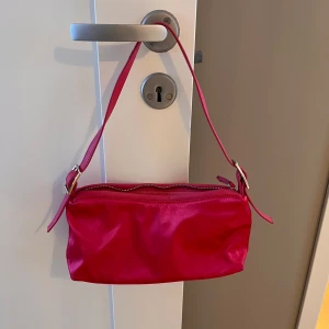 Rosa axelväska med dragkedja - Snygg rosa axelväska i syntetmaterial med en praktisk dragkedja upptill. Perfekt för att bära med sig det viktigaste på stan eller till en utekväll. Enkel och stilren design som passar till många olika outfits. Använd 1 gång 