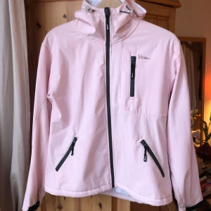 Rosa vindjacka  - Säljer en ljusrosa vindjacka från W med huva och dragkedja framtill. Jackan har tre fickor med dragkedjor och en logotyp på bröstet och ärmen. Perfekt för blåsiga dagar!