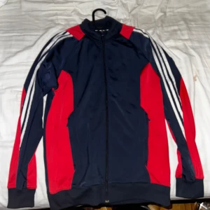Blå och röd jacka från Adidas - Säljer en snygg blå och röd jacka från Adidas med dragkedja och vita ränder längs ärmarna. Perfekt för en sportig look. Jackan har långa ärmar och ribbade muddar vid ärmslut och nederkant.