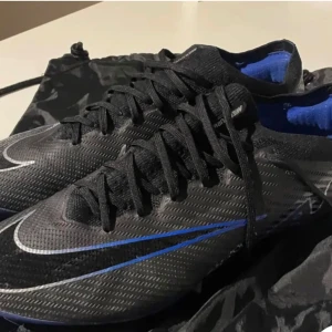 Nike Mercurial fotbollsskor endast använda 2 gånger!!! - Snygga svarta Nike Mercurial fotbollsskor med blå och vita detaljer. Skorna har snörning och är perfekta för fotbollsplanen. De kommer med en praktisk svart förvaringspåse.