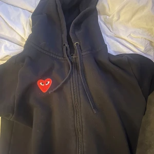 Cdg tröja  - Säljer en svart cdg hoodie med dragkedja och ett broderat rött hjärta med ögon på bröstet. Perfekt för en avslappnad stil.