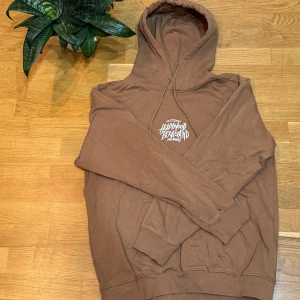 Brun hoodie med tryck - Säljer en brun hoodie från Hollywood Boulevard med tryck på både framsidan och baksidan. Hoodien har en stor ficka framtill och en bekväm huva. Perfekt för en avslappnad stil.