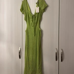 Grön glittrig klänning från Bershka - Snygg grön klänning från Bershka med glittrigt mönster. Den har en v-ringad design med knytning framtill och korta ärmar. Perfekt för en kväll ute!