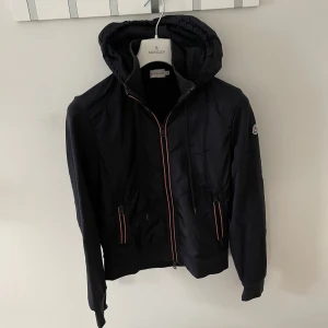 Svart jacka från Moncler - Snygg svart jacka från Moncler med huva och dragkedja. Jackan har långa ärmar och ribbade muddar. Perfekt för en stilren look. Skick 9/10, passar S. 
