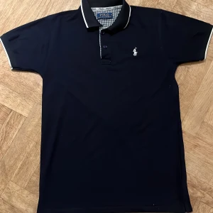 Marinblå pikétröja från Ralph Lauren - Snygg marinblå pikétröja från Ralph Lauren med klassisk krage och knappar. Tröjan har en broderad logotyp på bröstet och vita detaljer på kragen och ärmsluten. Använd fåtalgånger i bra skick. Pris kan diskuteras!