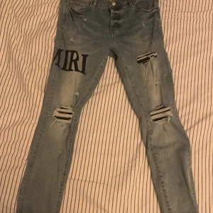 Blå jeans med slitningar från Amiri - Snygga blå jeans från Amiri med slitningar och tryck på ena benet. De har en modern slim passform och är perfekta för en trendig look. Midjan är midwaist och de har klassiska fem fickor samt knappgylf.