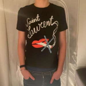 Svart t-shirt från Saint Laurent - Snygg svart t-shirt från Saint Laurent med ett coolt tryck av röda läppar och texten 'Saint Laurent' i vitt. Perfekt för en stilren look med en twist. Väldigt svår att få tag i!