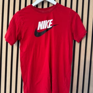 Röd t-shirt från Nike - Snygg röd t-shirt från Nike med det klassiska logotyptrycket i vitt och svart på bröstet. Perfekt för en sportig look eller avslappnad stil.