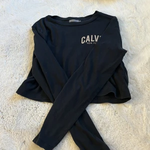 Svart långärmad tröja från Calvin Klein - Säljer en svart långärmad tröja från Calvin Klein med tryck 'CALVIN NEW YORK' på bröstet. Perfekt för en stilren och avslappnad look. Tröjan är i storlek M och har en klassisk passform.