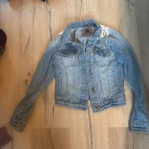 Jeansjacka med spetsdetalj från Lab Industries - Snygg jeansjacka från Lab Industries med unik spetsdetalj på ryggen. Jackan har knappar framtill och två bröstfickor. Perfekt för att ge din outfit en cool touch.