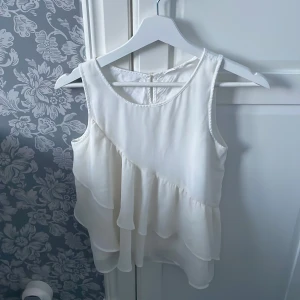 Vit ärmlös blus med volanger - Säljer en söt vit blus med volanger och knappdetalj i ryggen. Den är ärmlös och har en luftig design som passar perfekt för varmare dagar. Perfekt för en stilren och elegant look.