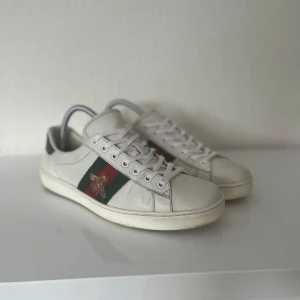 Gucci ace  - Snygga vita sneakers från Gucci med broderad bi-detalj på sidan. Skorna har klassiska gröna och röda ränder samt metallic-detaljer i rött och grönt på hälen. Perfekta för en stilren look. Väldigt bra skick. Tveka inte om du har någon fråga.