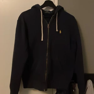 Mörkblå zip hoodie från Ralph Lauren - Säljer en stilren mörkblå hoodie från Ralph Lauren med dragkedja och den klassiska broderade loggan i gult på bröstet. Perfekt för en avslappnad look. Storlek S men passar också XS. Skick 8/10.