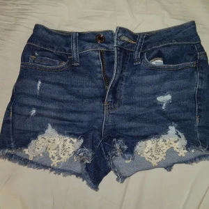 Blå jeansshorts med spetsdetaljer - Snygga blå jeansshorts från SO med slitna detaljer och vit spets vid benen. De är högmidjade och har en klassisk femficksdesign. Perfekta för en avslappnad sommarlook!