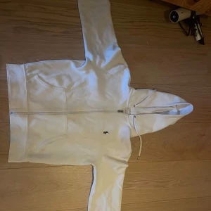 Vit hoodie från Ralph Lauren - Säljer en stilren vit hoodie från Ralph Lauren med dragkedja och huva. Den har en liten broderad logga på bröstet och praktiska fickor framtill. Perfekt för en avslappnad look. Storleken är S, 170/92A. Köpt från zalando för 1695kr
