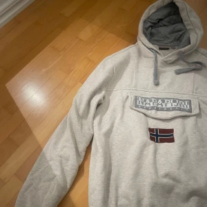 Grå hoodie från Napapijri - Säljer en stilren grå hoodie från Napapijri med en stor logotyp och norsk flagga på framsidan. Tröjan har en justerbar huva med snören och långa ärmar. Perfekt för kyliga dagar.