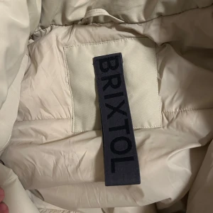 Beige pufferjacka från Brixtol - Snygg beige pufferjacka från Brixtol med huva och dragkedja. Jackan har praktiska fickor och är perfekt för kyligare dagar. Tillverkad i 100% polyester för både foder och yttertyg.