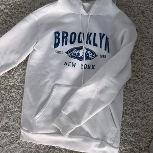 Vit hoodie med Brooklyn-tryck - Snygg vit hoodie med blått Brooklyn-tryck. Lite nopprig nertill men annars i bra skick. Storlek S