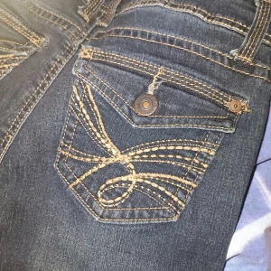 Mörkblå jeans med broderade detaljer - Snygga mörkblå jeans med unika broderade detaljer på bakfickan. Perfekta för en stilren look med en twist. Klassisk design med knappar och dragkedja.