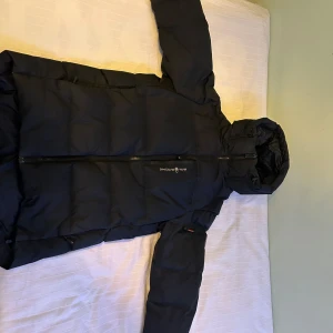 Svart quiltad från Sail Racing - Snygg och varm svart quiltad från Sail Racing (Sail Antarctica Expedition) med huva och dragkedja. Perfekt för kalla vinterdagar. Jackan har en stilren design med långa ärmar och är idealisk för att hålla värmen. Pris kan diskuteras vid snabb affär😀