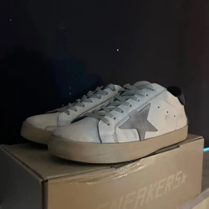 Vita sneakers från Golden Goose - Snygga vita sneakers från Golden Goose med en grå stjärndetalj på sidan. Skorna har en blå häl och är tillverkade i skinn med snörning framtill. Perfekta för en avslappnad stil.