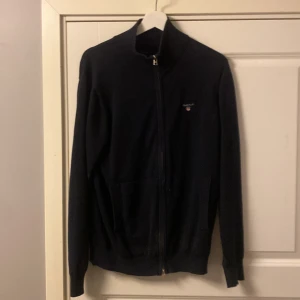 Svart kofta från Gant - Snygg svart kofta från Gant med dragkedja framtill. Perfekt för kyligare dagar och har en stilren design med diskret logga på bröstet. Passar bra till både vardag och mer uppklädda tillfällen.