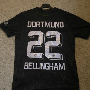 Fotbolls tröja - Säljer en svart Dortmund fotbollströja från Puma med nummer 22 och namnet Bellingham på ryggen. Tröjan har korta ärmar och klubbens emblem på bröstet tillsammans med sponsorlogga. Perfekt för fans av laget! Den är också i väldigt fint skick och är storlek M.