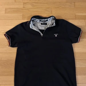 Svart kortärmad pikétröja från Gant - Snygg svart pikétröja från Gant med korta ärmar och randiga detaljer på kragen och ärmsluten. Perfekt för en stilren look. Klassisk design med broderad logotyp på bröstet.