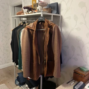 Coloniaire jacka - Snygg brun kappa med huva. Jackan har en kombination av mocka och ett mönstrat tyg, vilket ger den en unik look. Den har knappar framtill och praktiska fickor. Perfekt för höst och vår. Den är i storlek 50 (M-L). Nypris ligger på 6999kr. Är endast använd 4 gånger!. Skriv gärna om ni har frågor🤗