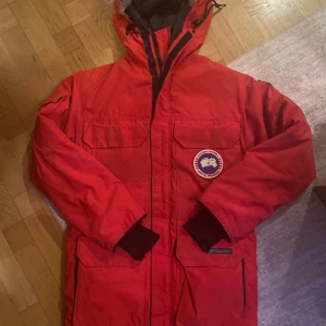 Röd parkajacka från Canada Goose - Säljer en röd parkajacka från Canada Goose med ikonisk logga på bröstet. Jackan har en huva och flera praktiska fickor. Perfekt för kalla vinterdagar.