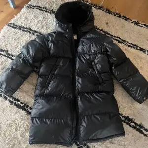 Säljer en svart dunjacka från Nike med Therma-Fit teknologi. Jackan har en hög krage och dragkedja framtill. Perfekt för kalla dagar med sin långa passform och vadderade design.