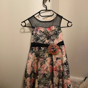 Blommig klänning från Miss Seviva - Charmig blommig klänning från Miss Seviva med ett svart meshok och ett dekorativt bälte med blomdetalj i midjan. Klänningen har en vacker blommönster i rosa och gröna toner och en dragkedja i ryggen.