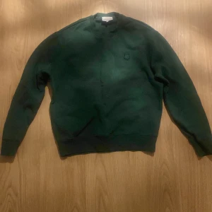 Grön tröja från Maison Kitsuné - Säljer en stilren grön tröja från Maison Kitsuné med ett broderat emblem på bröstet. Tröjan har långa ärmar och en klassisk rund halsringning. Perfekt för en avslappnad look.