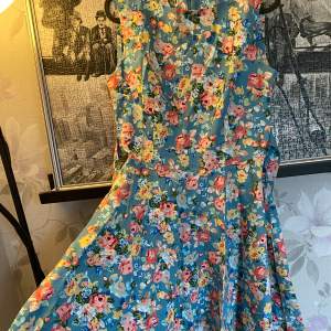 Charmig blå klänning med ett färgglatt blommönster. Den har en ärmlös design och en vacker passform som ger en feminin siluett. Perfekt för somriga dagar.