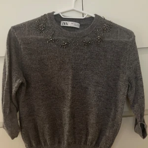 Grå tröja med pärldetaljer från Zara - Säljer en stilren grå tröja från Zara med vackra pärldetaljer runt halsringningen. Tröjan har 3/4 ärmar och tunt fint stickad . Ett måste för garderoben! 🖤