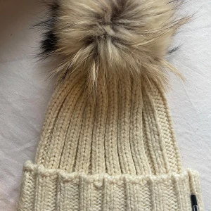 Beige stickad mössa med pälsboll - Säljer en vit/beige stickad mössa med en stor pälsboll på toppen. Äkta päls