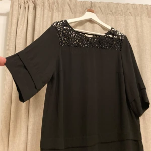 Svart blus från Monsoon - Elegant svart blus från Monsoon med vackra spetsdetaljer vid halsringningen. Blusen har trekvartsärmar och en lös passform, perfekt för en stilren look.