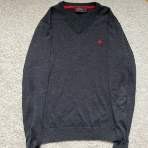 Grå tröja i merinoull från Morris - Säljer en stilren grå tröja från Morris i 100% merinoull. Tröjan har en v-ringad design och ett rött broderat emblem på bröstet. Perfekt för en elegant och bekväm look.