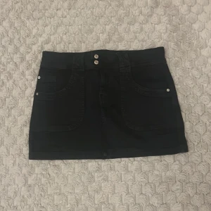 Svart kortkjol från Bershka - Snygg svart kortkjol från Bershka med två knappar framtill och praktiska fickor både fram och bak. Perfekt för en stilren look.