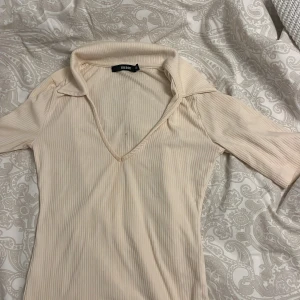 Beige ribbad polotopp från Bik Bok - Säljer en stilren beige ribbad polotopp från Bik Bok. Toppen har en djup v-ringning och korta ärmar, perfekt för en chic look. Passar bra till både jeans och kjol.