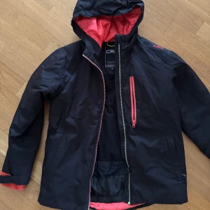 Svart vindjacka från CMP - Säljer en svart vindjacka från CMP med röd dragkedja och huva. Jackan har en quiltad insida och en bröstficka med dragkedja. Perfekt för blåsiga dagar!