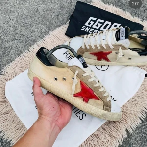 Golden Goose sneakers med röd stjärna - Snygga Golden Goose sneakers med en iögonfallande röd stjärna på sidan. Skorna har en beige och vit bas med svarta detaljer vid hälen. De är utrustade med snörning och har en klassisk sneakerdesign.