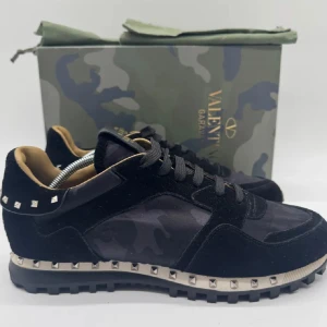 Svarta sneakers från Valentino - Snygga svarta sneakers från Valentino med coola nitar runt sulan och ett diskret camouflage-mönster. Skorna har snörning och en stilren design som passar perfekt för en trendig look.