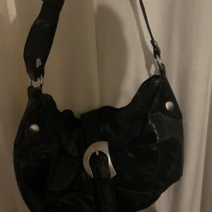 Svart axelväska med spänne - Snygg svart axelväska med ett stort silverfärgat spänne framtill. Väskan har en justerbar axelrem och dekorativa nitar. Perfekt för att bära dina dagliga nödvändigheter med stil.