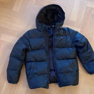Svart dunjacka från Tommy Hilfiger - Säljer en snygg svart dunjacka från Tommy Hilfiger med huva och dragkedja. Jackan har ett diskret märke på bröstet och är perfekt för kyligare dagar. Den är vadderad och ger en skön värme. Jackan är använd en vintersäsong. Inga skador eller fläckar återges. 