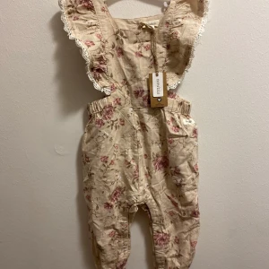 Blommig byxdress från Newbie - Söt blommig byxdress från Newbie i beige med rosa blommor. Byxdressen har volangdetaljer och spetskant vid axlarna. Perfekt för små barn i åldern 9-12 k.