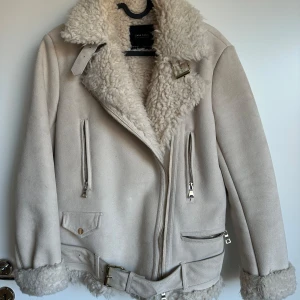 Beige jacka ZARA - Säljer en snygg beige jacka från Zara med fuskpälsfoder och krage. Jackan har dragkedja, bälte i midjan och flera fickor med dragkedjor. Perfekt för kyliga dagar och ger en stilren look. Storlek S