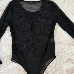 Svart långärmad body från In Vogue Paris - Snygg svart body med långärmade meshärmar och blommiga spetsdetaljer framtill. Den har en rund halsringning och är tillverkad i ett lätt och bekvämt material. Storlek L men skulle säga att den passar M också även en liten M. Perfekt till fest